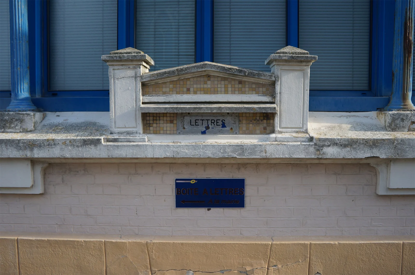 Ancienne fente de boîte aux lettres marquée « Lettres » dans un muret de pierre, sous une fenêtre bleue